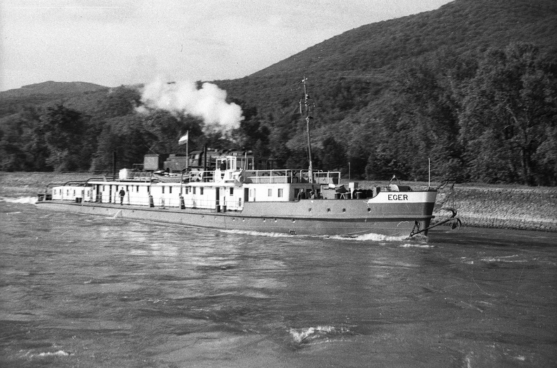 a Vaskapu-szoros hajózócsatornájában az Eger vontatóhajó, mögötte a Duna-partján egy vontató gőzmozdony.